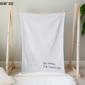 May include: A white throw blanket with the text "go away, I'm reading." printed in black on a wooden stand.