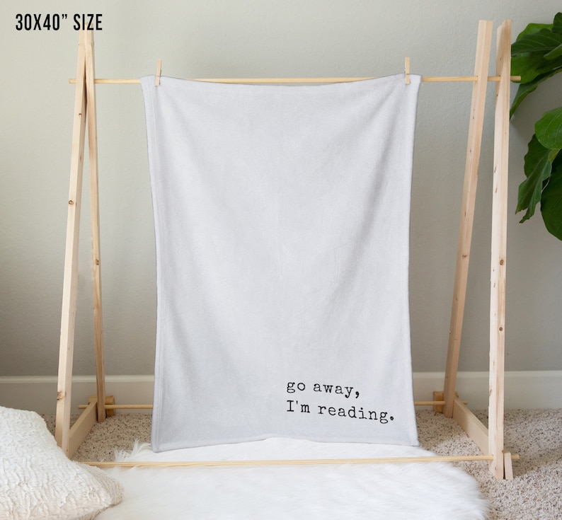 May include: A white throw blanket with the text "go away, I'm reading." printed in black on a wooden stand.
