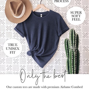 May include: A navy blue t-shirt hanging on a wooden hanger against a white background with a black and white geometric pattern. The t-shirt is made with premium Airlume Combed and Ring-Spun Cotton. The text "ECO-FRIENDLY PROCESS", "SUPER SOFT FEEL", and "TRUE UNISEX FIT" are visible in white circles. The text "Only the best" is written in black cursive.