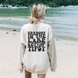 May include: Cream-colored sweatshirt with the text "ERRBODY IN THE LAKE GETTIN' TIPSY" in black, retro-style lettering. The sweatshirt is worn on a beach with the ocean in the background. A casual, relaxed style.