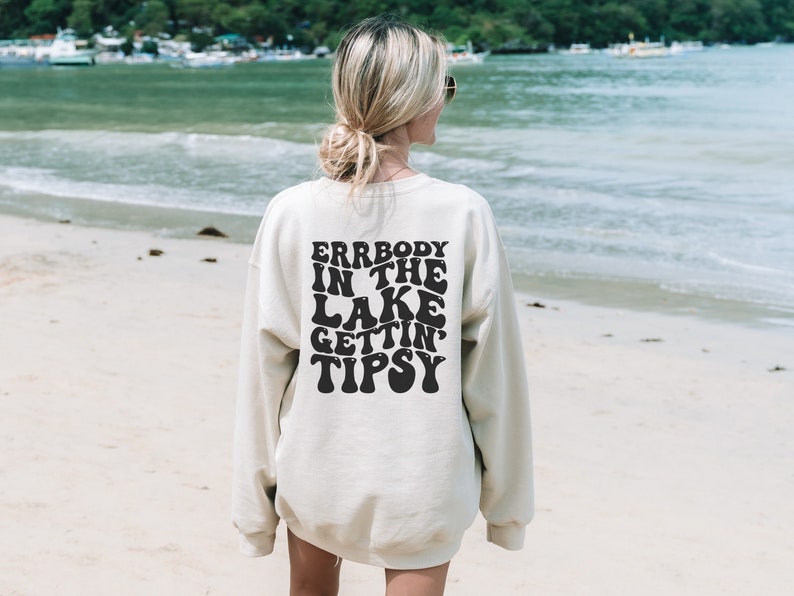 May include: Cream-colored sweatshirt with the text "ERRBODY IN THE LAKE GETTIN' TIPSY" in black, retro-style lettering. The sweatshirt is worn on a beach with the ocean in the background. A casual, relaxed style.