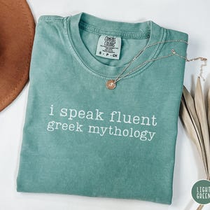 May include: A light green t-shirt with the text "i speak fluent Greek mythology" in white. The shirt is folded and has a silver necklace with a pendant. A brown hat and dried leaves are in the background.