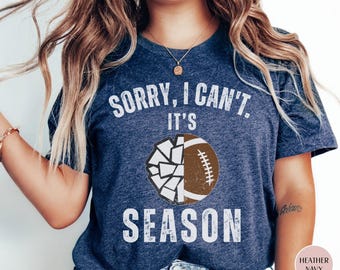 May include: Heather navy t-shirt with the text "SORRY, I CAN'T, IT'S SEASON" in white. A graphic of a football, half shattered, is centered below the text. The shirt is a casual style.