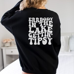 May include: Black sweatshirt with white text on the back that reads "ERRBODY IN THE LAKE GETTIN' TIPSY" in a retro font. The sweatshirt is a solid black color and has long sleeves.
