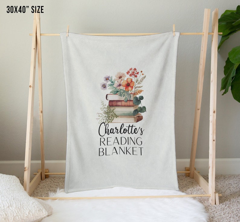 May include: A white fleece blanket with a stack of books and flowers printed on it. The text "Charlotte's Reading Blanket" is printed below the books.