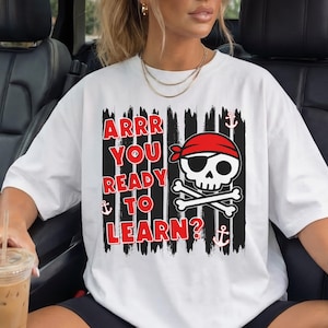 May include: A white short-sleeve t-shirt featuring a graphic print with black vertical brush strokes, red text that reads ARRR YOU READY TO LEARN?, a skull wearing a red bandana and eye patch, and small white anchor icons.