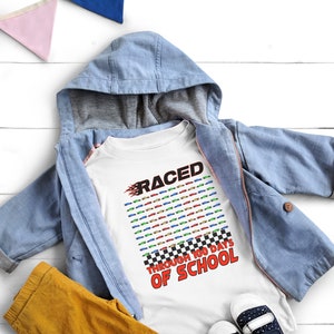 K&ouml;nnte beinhalten: Ein wei&szlig;es T-Shirt mit dem Text "RACED THROUGH 100 DAYS OF SCHOOL" in Rot und Schwarz. Das Shirt zeigt ein Design mit bunten Rennwagen. Eine hellblaue Kapuzenjacke und eine gelbe Hose liegen auf einer wei&szlig;en Holzoberfl&auml;che.