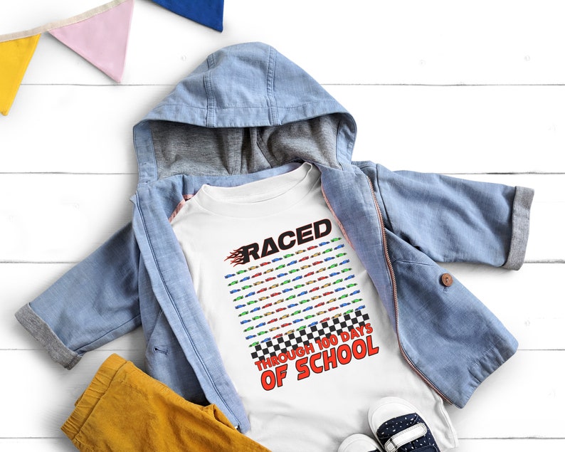 K&ouml;nnte beinhalten: Ein wei&szlig;es T-Shirt mit dem Text "RACED THROUGH 100 DAYS OF SCHOOL" in Rot und Schwarz. Das Shirt zeigt ein Design mit bunten Rennwagen. Eine hellblaue Kapuzenjacke und eine gelbe Hose liegen auf einer wei&szlig;en Holzoberfl&auml;che.