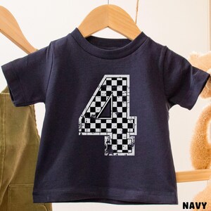 Pu&ograve; includere: T-shirt blu navy per bambini con un grande numero "4" in un design a bandiera a scacchi. Il numero &egrave; bianco con un motivo a scacchi bianco e nero. La maglietta &egrave; appesa a un appendiabiti di legno.