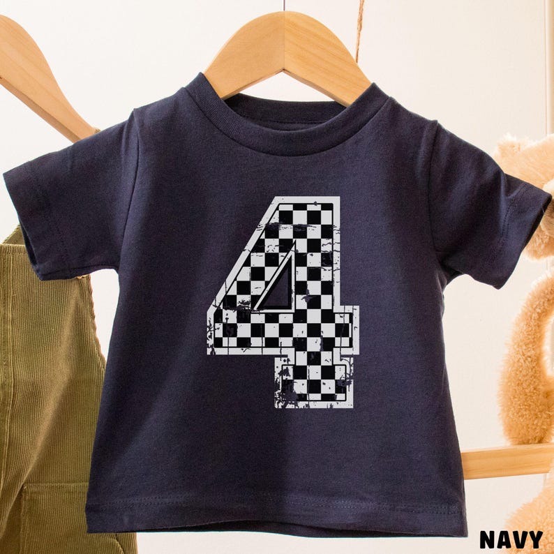 Pu&ograve; includere: T-shirt blu navy per bambini con un grande numero "4" in un design a bandiera a scacchi. Il numero &egrave; bianco con un motivo a scacchi bianco e nero. La maglietta &egrave; appesa a un appendiabiti di legno.