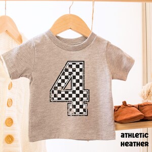 Pu&ograve; includere: T-shirt grigio atletico per bambini piccoli con un grande numero 4 a scacchi bianchi e neri. La maglietta &egrave; appesa a un appendiabiti di legno. Il testo "ATHLETIC HEATHER" &egrave; stampato in nero.