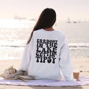 May include: A light gray sweatshirt with the text "ERRBODY IN THE LAKE GETTIN' TIPSY" in black, retro-style lettering. The sweatshirt is worn on a person sitting on a beach. A bouquet of flowers and a book are visible.