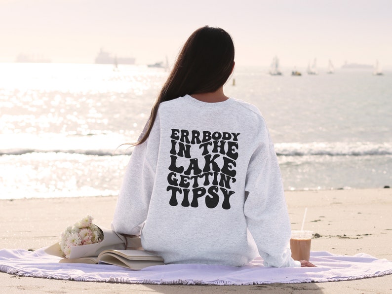 May include: A light gray sweatshirt with the text "ERRBODY IN THE LAKE GETTIN' TIPSY" in black, retro-style lettering. The sweatshirt is worn on a person sitting on a beach. A bouquet of flowers and a book are visible.
