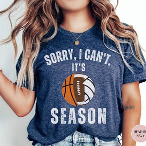 May include: Heather navy blue t-shirt with the text "SORRY, I CAN'T, IT'S SEASON" in white. The design features a basketball, a football, and a volleyball. The shirt is a casual wear item.