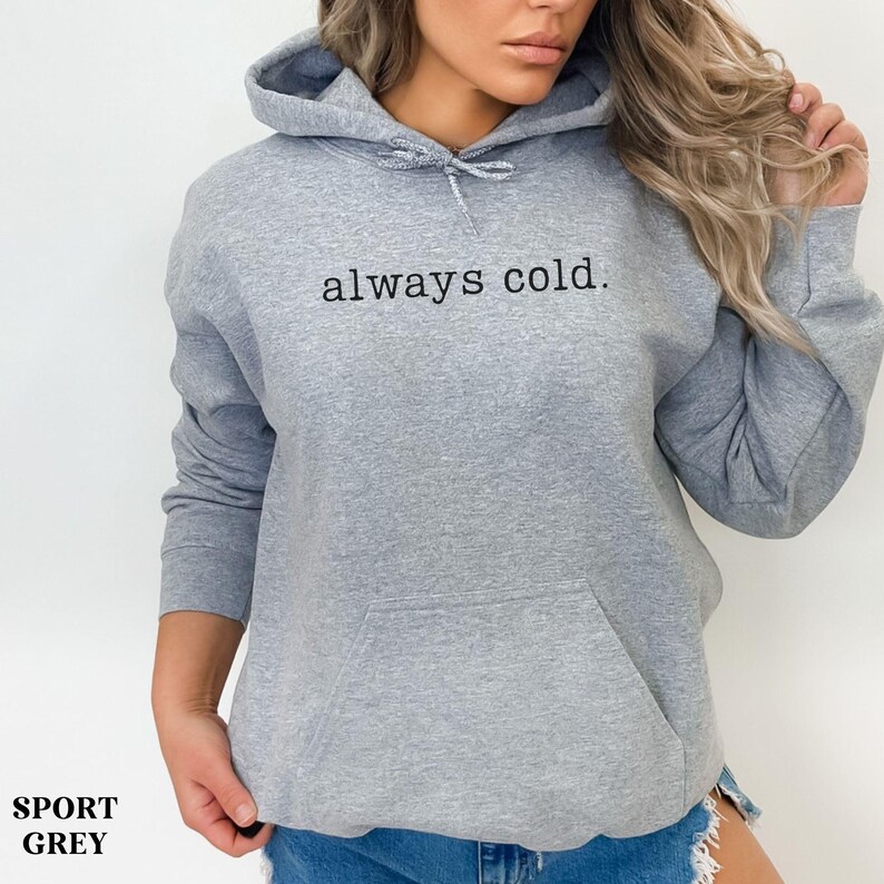 May include: A light grey hoodie with a drawstring hood and a front pocket. The text "always cold." is printed in black across the chest. The bottom left corner of the image has the text "SPORT GREY".