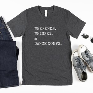 May include: A dark heather grey t-shirt with the text "WEEKENDS. WHISKEY. & DANCE COMPS." printed in white.