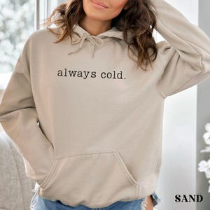 May include: Sand-colored hoodie with a drawstring hood and a front pocket. The phrase "always cold" is printed in black across the chest. The hoodie is made of a soft material.