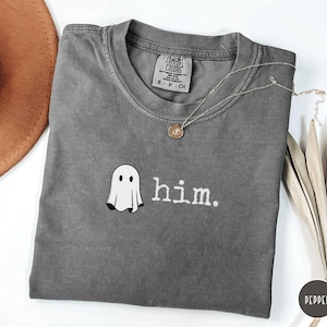 May include: A charcoal gray short-sleeve t-shirt featuring a small white ghost graphic and the lowercase text "him." printed in white. The shirt has a crew neckline and a Comfort Colors brand label on the inside collar indicating a size small.