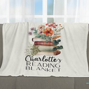 May include: A white fleece blanket with a floral design and a stack of books. The text "Charlotte's Reading Blanket" is printed on the blanket.