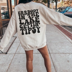 May include: Cream-colored sweatshirt with the text "Errbody in the Lake Gettin' Tipsy" in black, retro-style lettering. The sweatshirt is long-sleeved and worn with white sneakers. The image is taken outdoors.