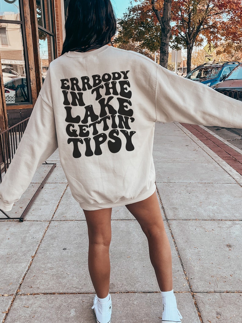 May include: Cream-colored sweatshirt with the text "Errbody in the Lake Gettin' Tipsy" in black, retro-style lettering. The sweatshirt is long-sleeved and worn with white sneakers. The image is taken outdoors.