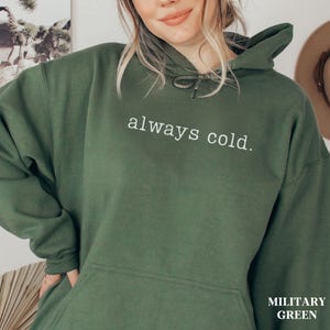 May include: Military green hoodie with the text "always cold." in white. The hoodie has a drawstring hood and a front pocket. The bottom right corner of the image has the text "MILITARY GREEN."