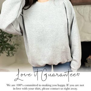 May include: A light gray sweatshirt with a crew neck. The text "Love it Guarantee" is written in cursive script. The sweatshirt is made of a soft material. The image is of a person wearing the sweatshirt.