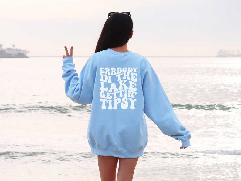 May include: A light blue sweatshirt with the text "ERRBODY IN THE LAKE GETTIN' TIPSY" in white, retro-style lettering. The sweatshirt is seen from the back, with a person standing near the water.
