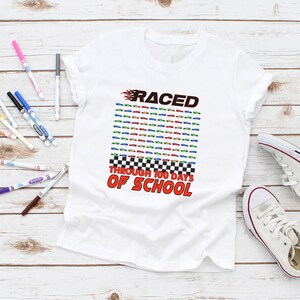 K&ouml;nnte beinhalten: Wei&szlig;es T-Shirt mit dem Text "RACED THROUGH 100 DAYS OF SCHOOL" in Rot und Schwarz. Das Design umfasst ein Raster aus bunten Rennwagen und ein Schachbrettmuster. Ein Paar wei&szlig;e Turnschuhe und bunte Marker sind ebenfalls im Bild.
