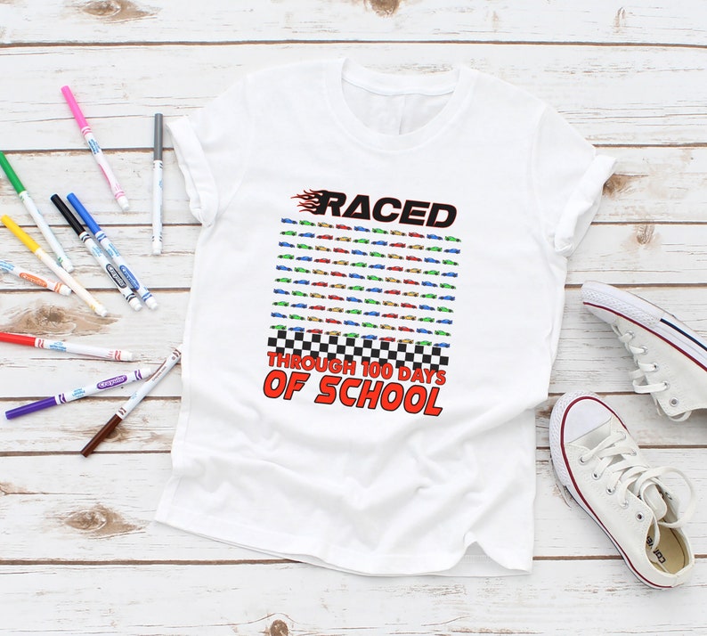 K&ouml;nnte beinhalten: Wei&szlig;es T-Shirt mit dem Text "RACED THROUGH 100 DAYS OF SCHOOL" in Rot und Schwarz. Das Design umfasst ein Raster aus bunten Rennwagen und ein Schachbrettmuster. Ein Paar wei&szlig;e Turnschuhe und bunte Marker sind ebenfalls im Bild.