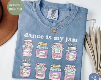 May include: Blue jean colored t-shirt with the phrase "dance is my jam" and illustrations of dance-themed jam jars. The jars are labeled with dance styles like Tap, Jazz, and Ballet. A gold necklace rests on the shirt.