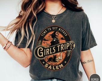 May include: Dark gray t-shirt with a circular graphic that reads "TIME TO GET WICKED GIRLS TRIP SALEM EST 1692 2026". The graphic features a witch, black cat, skull, and other Halloween-themed elements. The shirt is a comfortable, casual style.