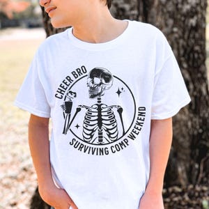 May include: A white short-sleeve t-shirt featuring a black graphic of a skeleton holding a drink inside a circle. The text reads CHEER BRO SURVIVING COMP WEEKEND. The design includes small star accents and is centered on the front of the garment.