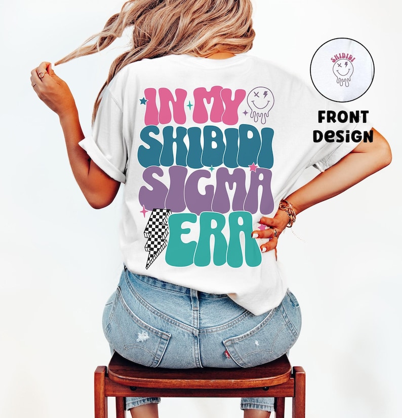 Puede incluir: Camiseta blanca con un dise&ntilde;o gr&aacute;fico que dice "In My Shibidi Sigma Era" en una fuente retro con un rayo y estrellas. El texto est&aacute; en tonos de rosa, morado, turquesa y verde.