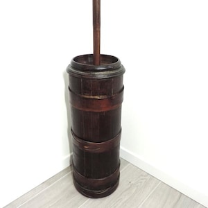 May include: A dark brown antique butter churn with a tall wooden handle. The churn is cylindrical, made of wooden staves held together by metal bands. The handle extends from the top.