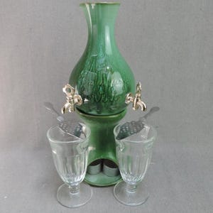 Peut inclure: Une fontaine à absinthe en céramique verte avec deux robinets en métal, portant le mot "Verte" en écriture cursive sur le devant. L'ensemble comprend deux verres à pied transparents et deux cuillères à absinthe perforées en métal.