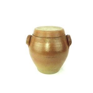 May include: A brown stoneware jar with a lid. The jar has a rounded body, two small handles, and horizontal ridges around the top and lid. The color gradient transitions from a light green at the base to a warm brown.