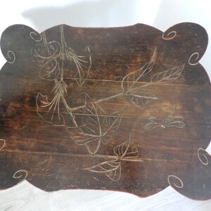 Puede incluir: Una mesa de madera de color marr&oacute;n oscuro con borde festoneado y superficie rectangular. La superficie presenta un dise&ntilde;o grabado de hojas, flores y una lib&eacute;lula, con patrones de espirales decorativos tallados en cada una de las cuatro esquinas.