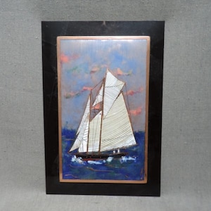 Może przedstawiać: Oprawione dzieło sztuki marynistycznej przedstawia żaglówkę z białymi żaglami na niebieskiej wodzie. Niebo ma pastelowe kolory. Grafika jest oprawiona w czarną prostokątną ramę z miedzianą obwódką, tworząc klasyczny motyw morski.