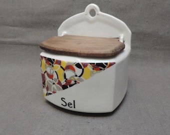 May include: White ceramic salt dispenser with a wooden lid and a geometric pattern in yellow, red, black, and gray. The dispenser has a hook on the back for hanging and says "Sel" on the front.