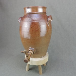 May include: A brown stoneware water crock with a spigot, lid, and two handles. The crock is on a small, round, wooden stand with three legs. The crock has a rustic, aged appearance.