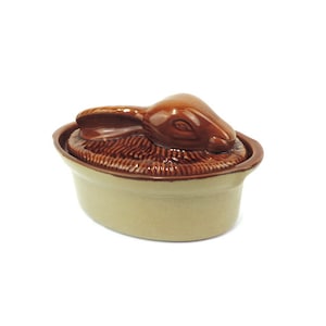 Op de afbeelding: Een keramische ovenschaal met een bruin konijnendeksel. De schaal is ovaalvormig en heeft een lichtbruine glazuur. Het konijnendeksel is gedetailleerd met een bruine glazuur en heeft een realistisch uiterlijk.