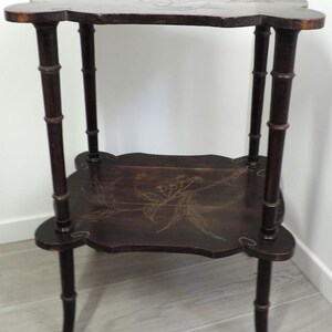 Puede incluir: Una mesa auxiliar de madera de dos niveles en color marr&oacute;n oscuro con cuatro patas dise&ntilde;adas para parecer tallos de bamb&uacute;. Tanto la parte superior cuadrada como el estante inferior tienen bordes festoneados y un patr&oacute;n floral incrustado.