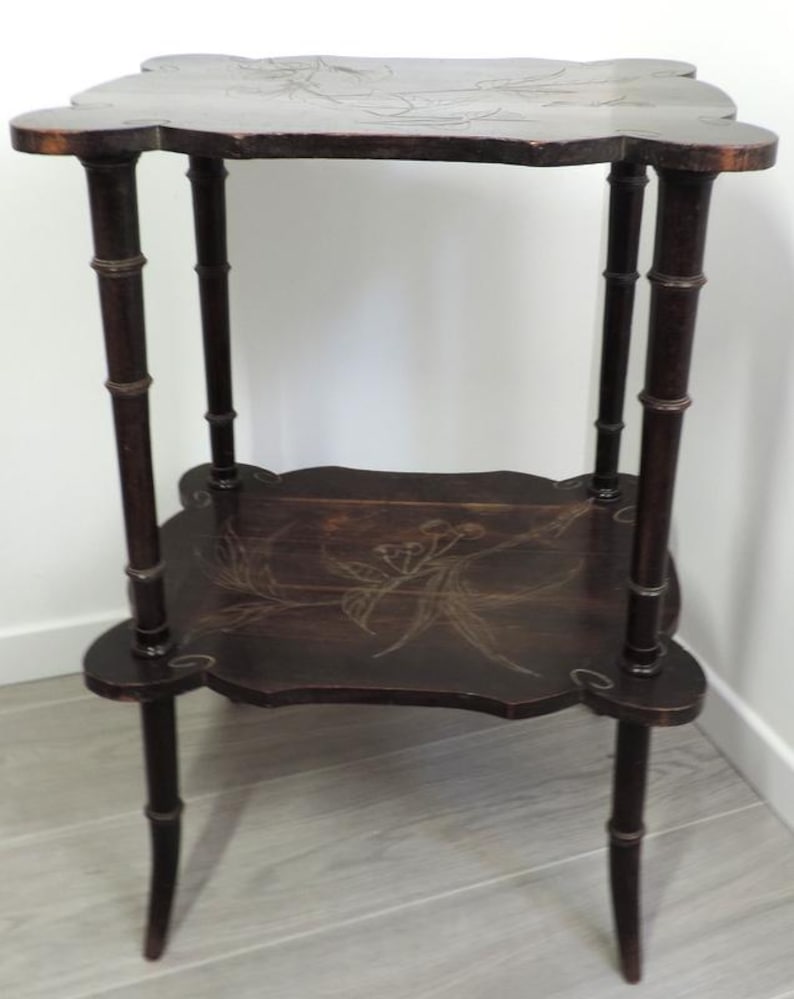 Puede incluir: Una mesa auxiliar de madera de dos niveles en color marr&oacute;n oscuro con cuatro patas dise&ntilde;adas para parecer tallos de bamb&uacute;. Tanto la parte superior cuadrada como el estante inferior tienen bordes festoneados y un patr&oacute;n floral incrustado.