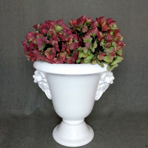 Puede incluir: Maceta de cerámica blanca con asas de cabeza de león, llena de un ramo de hortensias rosas y verdes.