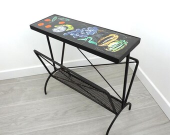 Puede incluir: Un revistero de metal negro con una parte superior rectangular con un diseño de frutas colorido. El revistero tiene un estante de malla y patas curvas. El diseño de frutas incluye uvas, naranjas y otras frutas.