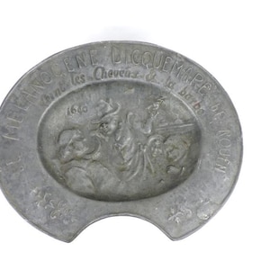 Op de afbeelding: Een zilveren metalen schotel met een reliëf van drie figuren en de tekst "LA MEGISSENE DICQUEMARE POUR LES CHEVAUX & LA BÊTE" rond de rand.