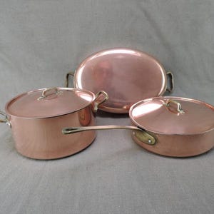 May include: Three copper cooking pots: a lidded pot, a lidded saucepan, and an oval serving dish. The copper pots have brass handles and lids. The cookware is displayed on a grey surface.
