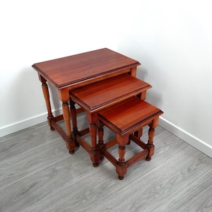 May include: A set of three nesting tables made of polished reddish-brown wood. Each table features a rectangular top with a smooth finish and four turned legs connected by lower support stretchers, arranged in a descending size order.