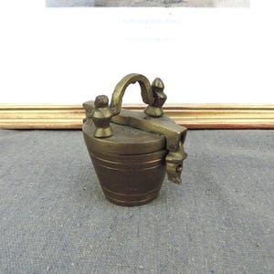 May include: An antique brass container with a handle and decorative features. The container has a cylindrical body with horizontal lines and a hinged lid. The handle is arched, and there are small, ornate figures on top. A framed print is in the background.
